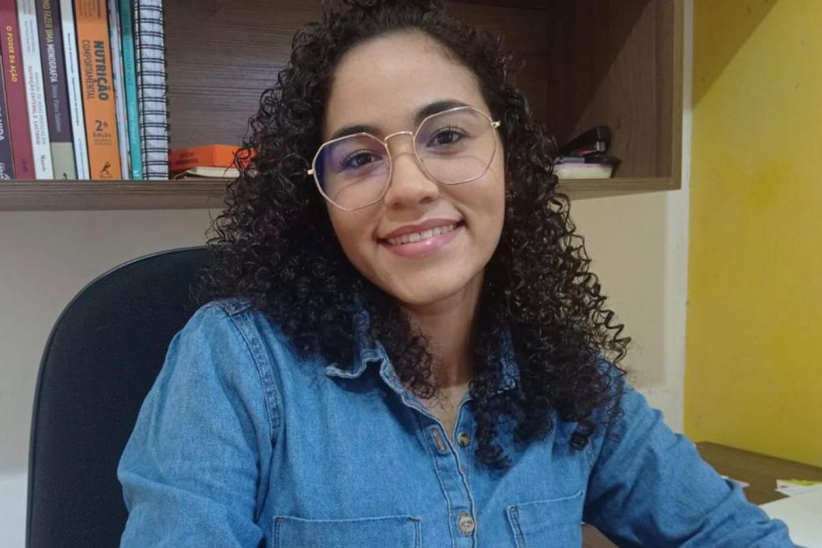 Nayara Monteiro em uma cadeira e uma estante com livros atrás